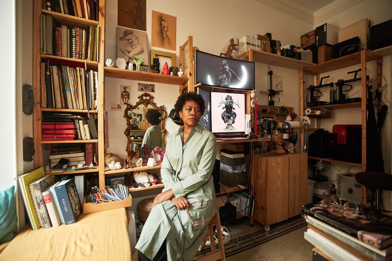 The artist in her studio, seated among shelves of books, sculptures and tools, with a digital work on the screen behind her.